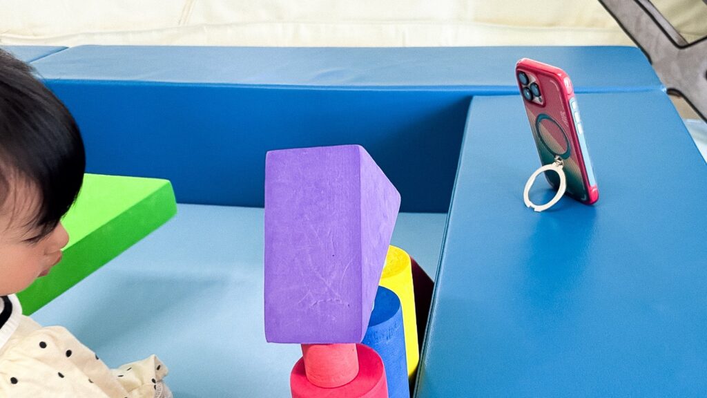キッズスペースでリングスタンドを使い縦向きに自立させたiPhoneケース。子どもが積み木で遊ぶ脇で動画を再生できる。