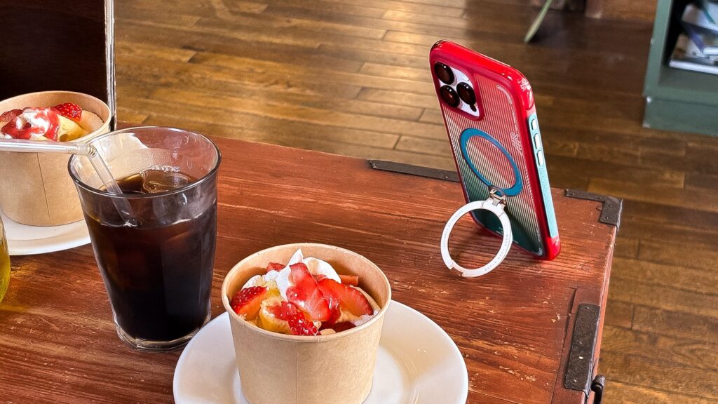 カフェのテーブルでリングスタンドを使い横向き自立させたケース。アイスコーヒーとフルーツボウルの隣で動画視聴に使える。