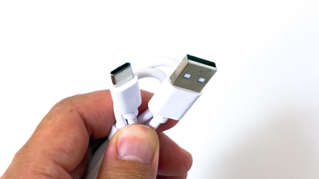付属の白いUSBケーブルを指で持って接写したもので、片端のUSB Type-Cコネクタと片端のUSB Type-Aコネクタが並び、短い充電用ケーブルであることが分かる。