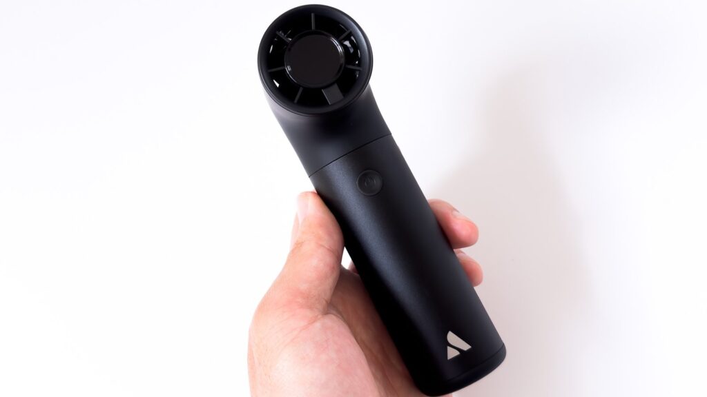 黒い円筒型ハンディファンを手のひらに乗せて斜めに構えた様子。上端の送風口にブレードと黒い円形パネル、握り部には電源ボタン、底面に三角形のロゴマークが見えている。