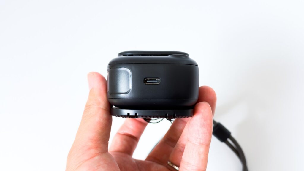 黒いネックファンの側面にあるUSB-C充電端子。本体を指でつまみ、丸い横長のポートが中央に見える。底面側には吸気口の格子も確認できる。