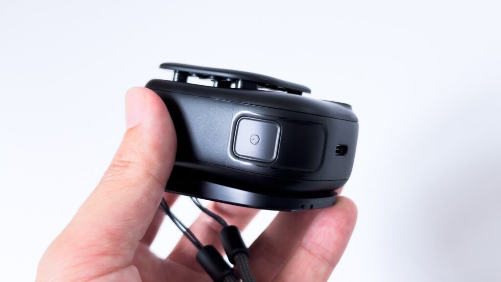 手に持った黒のネックファンの側面アップ。電源マークが刻まれた光沢のある四角いボタンが中央にあり、右隣には小さなストラップ取付穴が開いている。