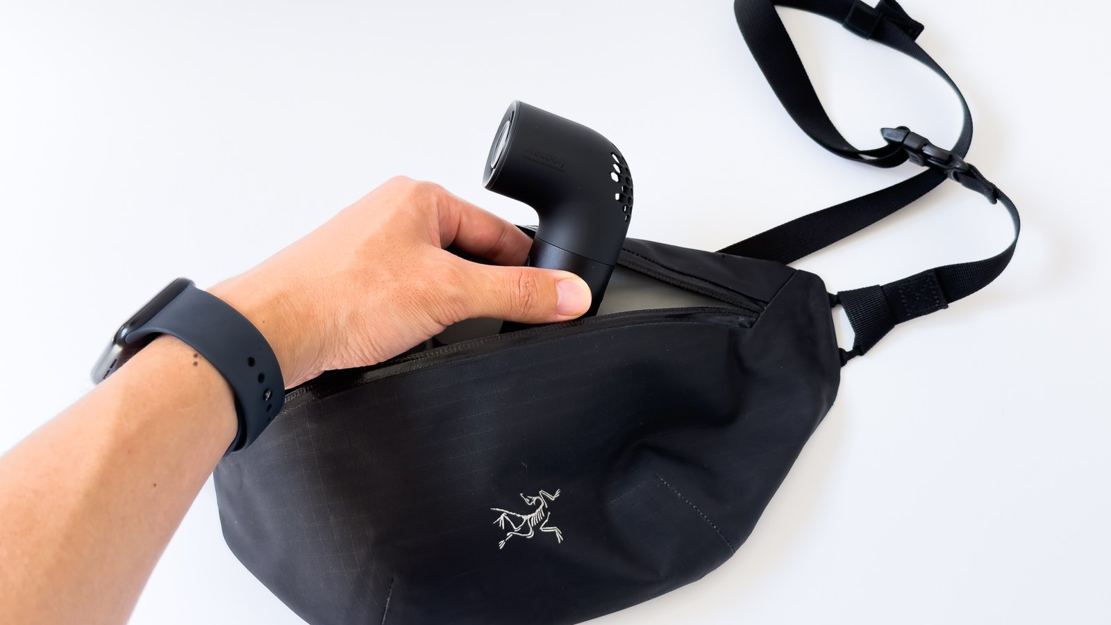 黒い四角いネックファンを黒のボディバッグへ収納する手元。斜めスリットの吹出口が上を向き、手首にはスマートウォッチが見える。