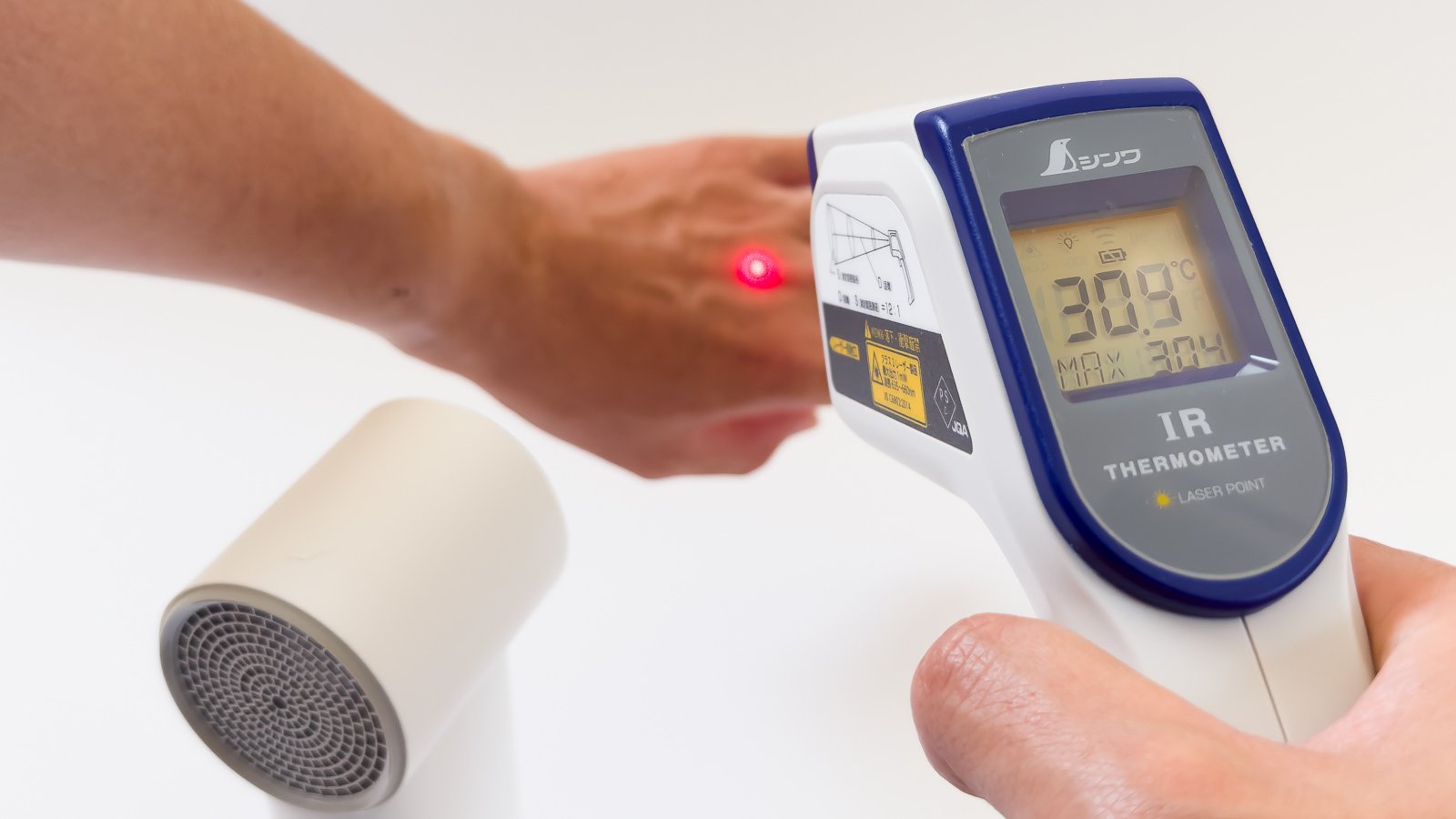 放射温度計を手の甲に当て、赤いレーザー点を照射しながら25.5℃を表示している様子。手前に白いミストファンの吹出口を上に向けて置き、冷却前後の差を計測している。