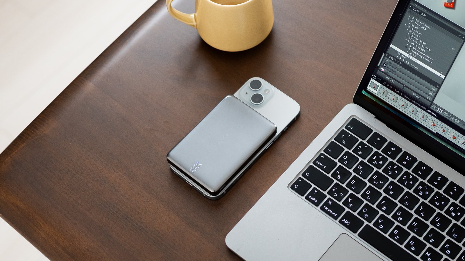 iPhoneに装着した本体を手に持つカット。背景の木目デスクにマウスとMacBookが置かれている。