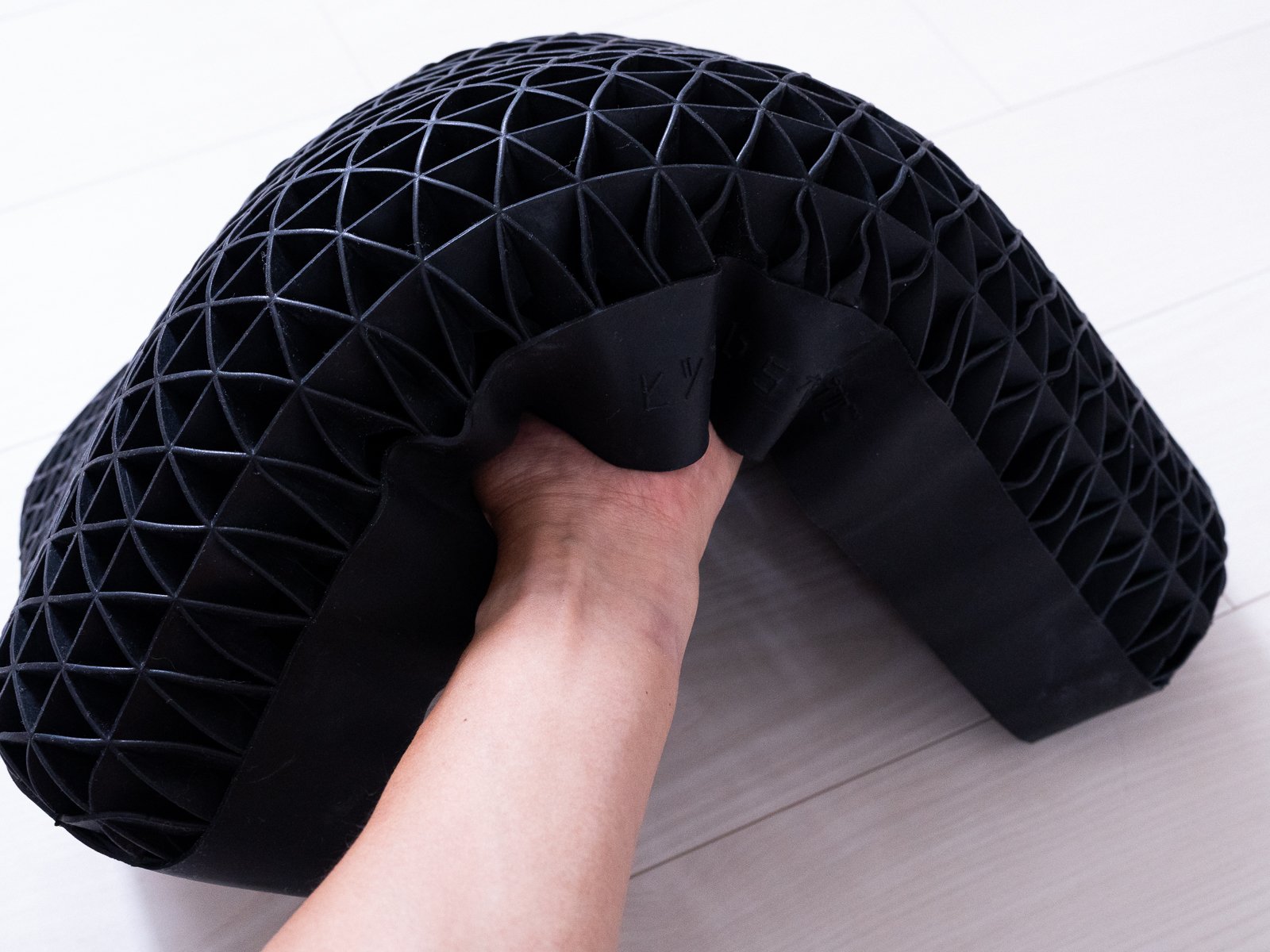 黒い本体を大きく折り曲げて片手で支えている様子。側面の帯には「ヒツジのいらない枕」の刻印が見え、折り曲げても裂けずに形が追従している。