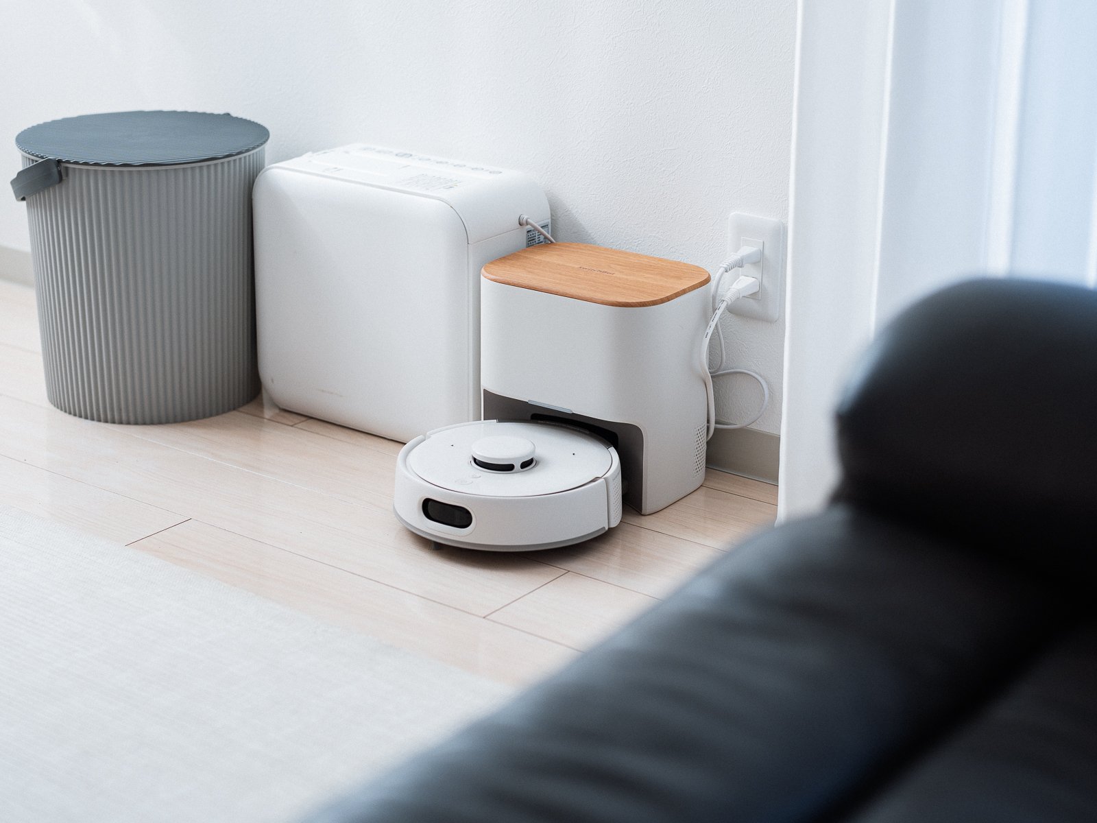 SwitchBot ロボット掃除機 K11+ Proがゴミ収集ステーションに戻っている様子。木目調のフタが付いた白いステーションがリビングの壁際に置かれている。
