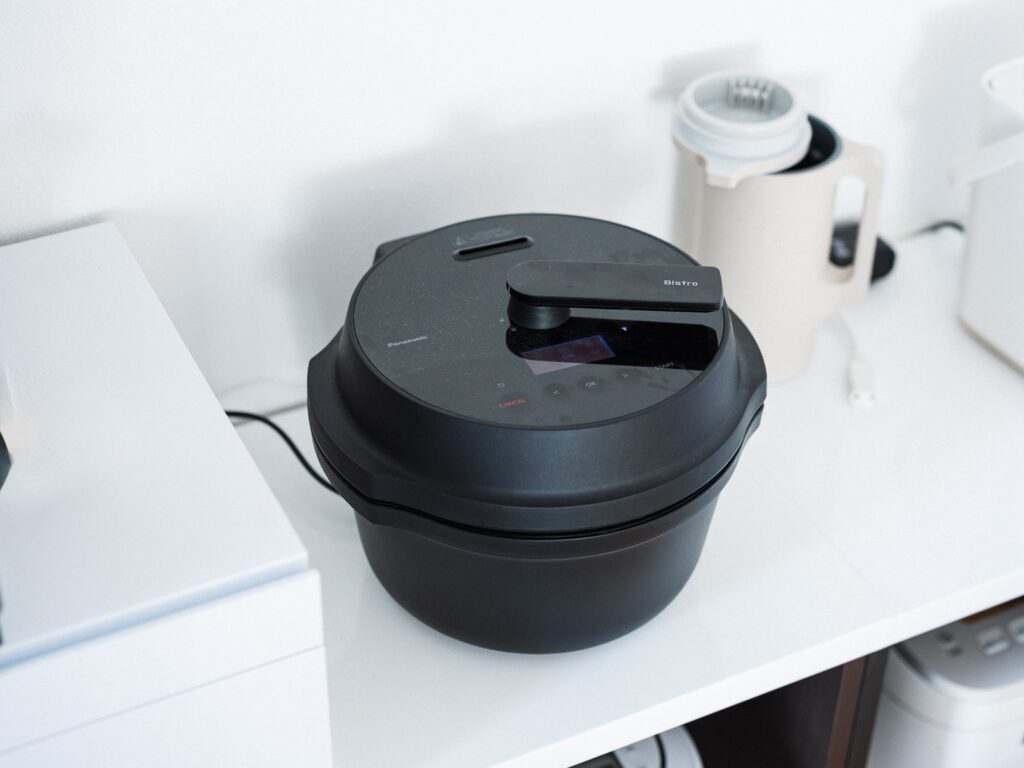 白い棚の上に置かれた黒いパナソニック オートクッカー NF-AC1000。ふたを閉じた状態で、奥に白いキッチン家電が並んでいる。