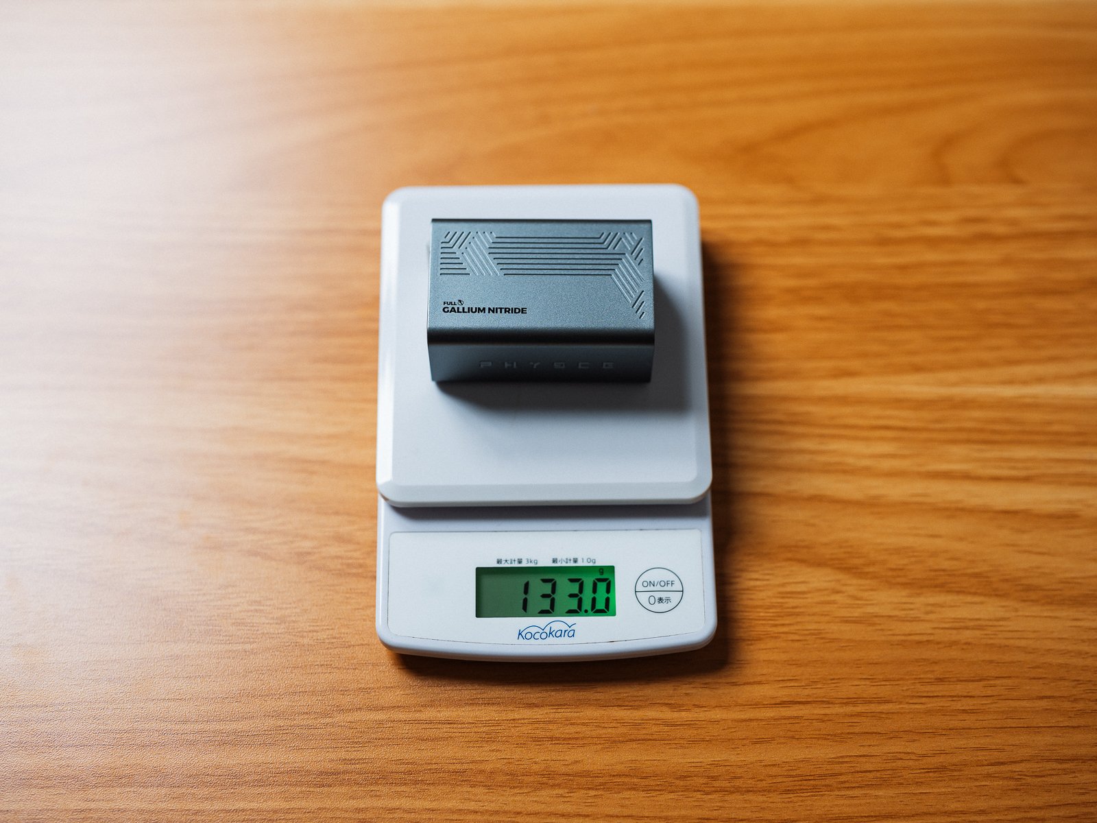 デジタルスケールに載せたPHYSCE 67W GaN急速充電器。液晶の表示は133.0gを示している。木目のデスクの上で計測している。