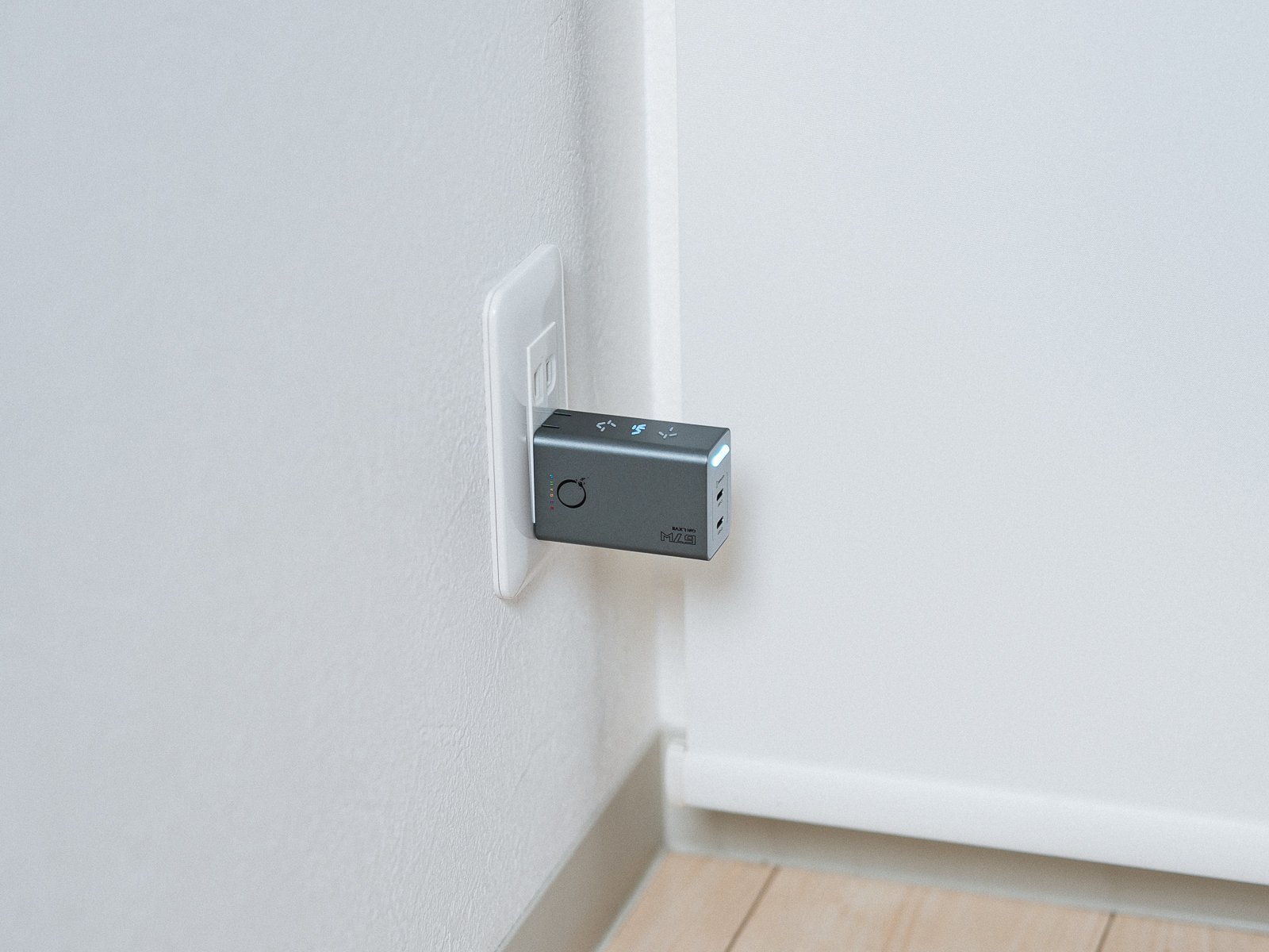 白い壁のコンセントに挿したPHYSCE 67W GaN急速充電器。床に近い位置のコンセントから、ウェッブグレーの本体が水平に突き出している。
