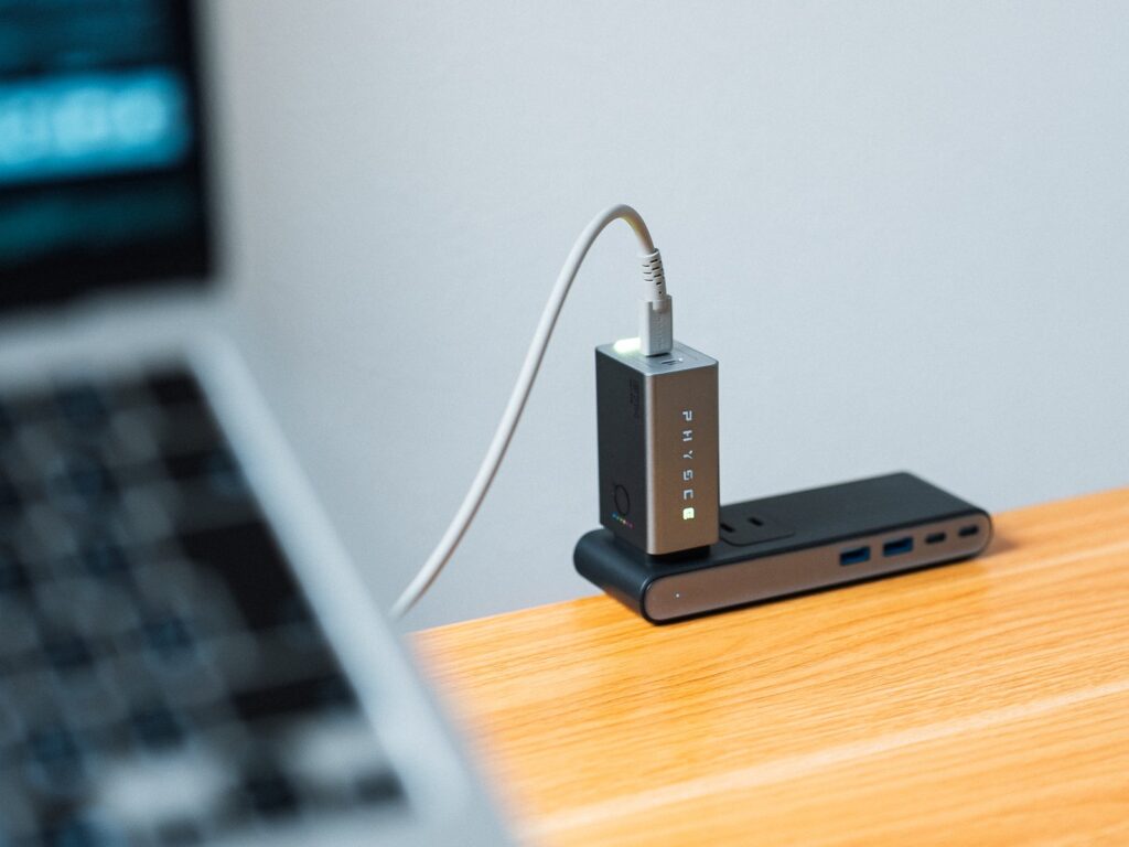電源タップに挿したPHYSCE 67W GaN急速充電器と、手前に置いたノートPC。本体側面のロゴの文字が見え、天面のポートからUSB-Cケーブルが伸びている。
