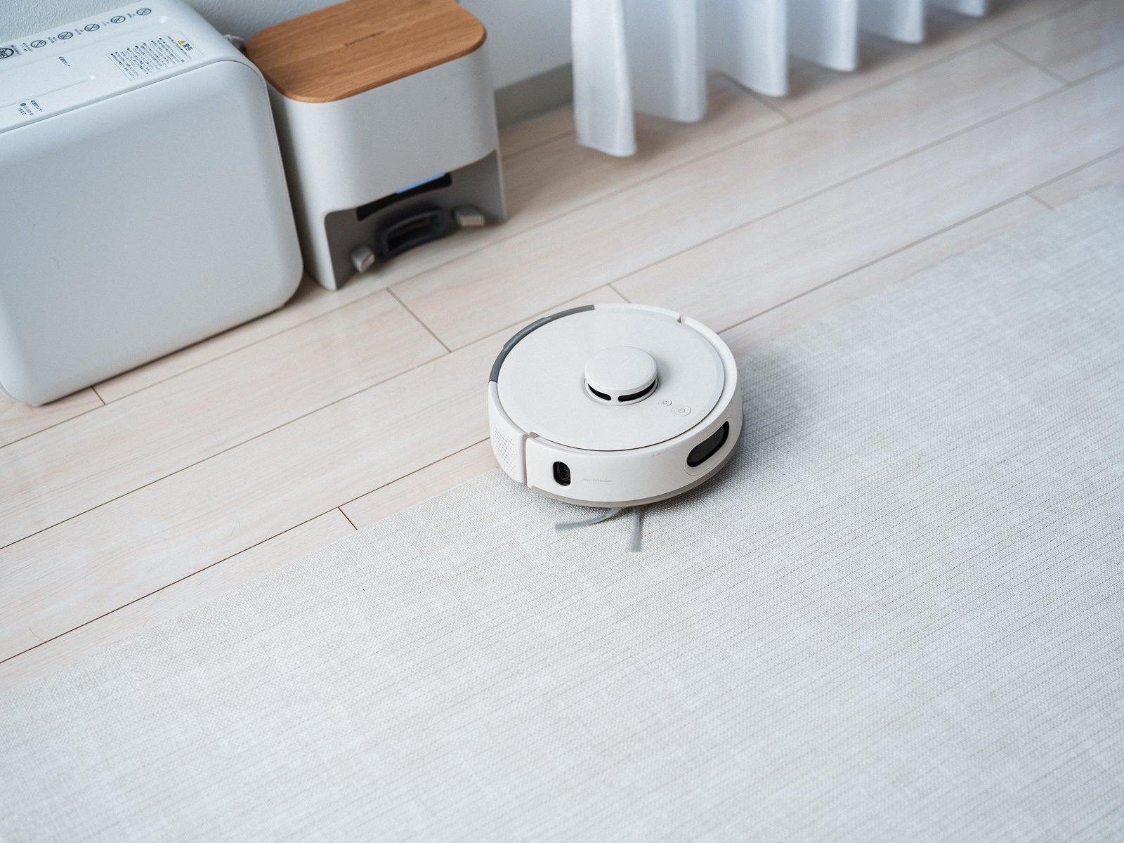 ラグの上を走行するSwitchBot ロボット掃除機 K11+ Pro。白い丸型の本体の奥の壁際に、ゴミを自動で吸い上げるステーションが置かれている。