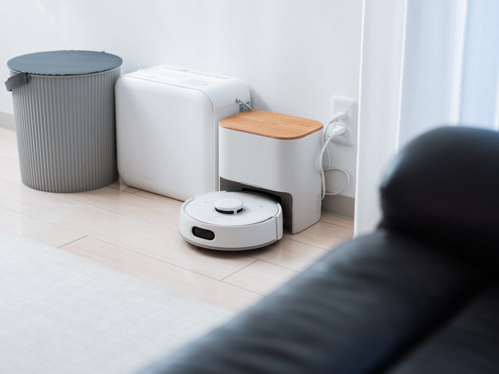 部屋の隅のステーションに戻ったSwitchBot ロボット掃除機 K11+ Pro。木目のふたが付いた白いステーションの横に、白い家電とグレーのゴミ箱が並ぶ。