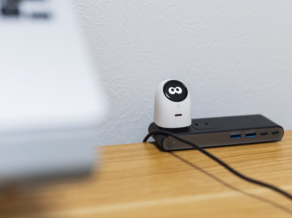白い壁を背景に、木製デスク上の黒い電源タップに挿したミルキー・ホワイトのTORRAS FlashEye Dot。丸い画面に白い目の表情が表示され、本体正面の下部にUSB-Cポートが見える。