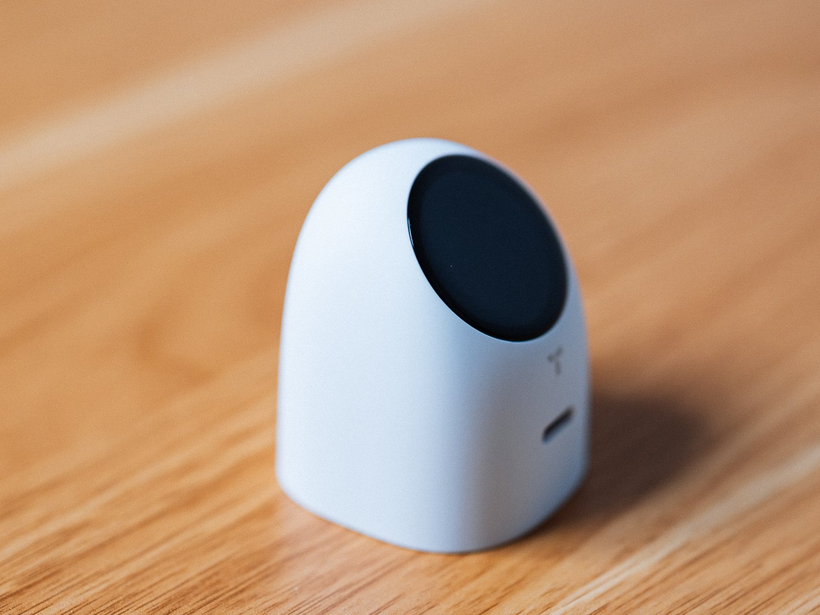 木製デスクに置いたTORRAS FlashEye Dotを斜め前から捉えた様子。角のない丸いボディで、上部の傾いた面に黒い円形のディスプレイがある。