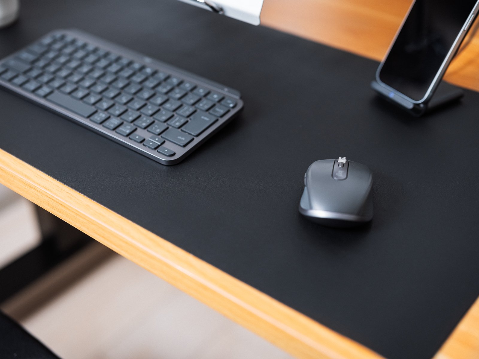黒いデスクマットの上にキーボードとマウス、スマホスタンドを並べた作業デスク。黒とグレーのガジェットでまとめられている様子が分かる。